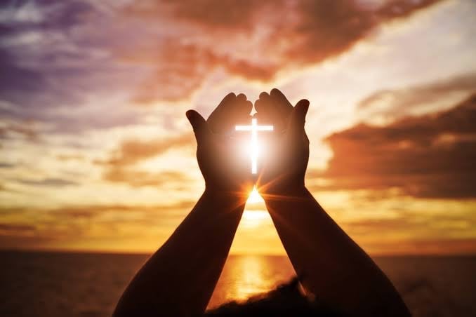 Hands lifted in prayer framing a glowing cross against a golden sunset sky